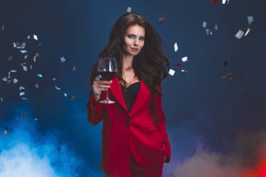 Parlak makyajlı, saç stili, kırmızı elbiseli, koyu arkaplanda bir cam ile poz veren bir kadın.