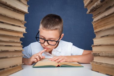 Gözlüklü zeki çocuk iki kitap yığınının arasında oturup kitap okuyor.