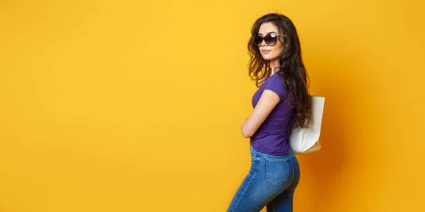 Güneş gözlüklü, mor gömlekli, kot pantolonlu, harika sarı arka planda çantasıyla poz veren güzel bir kadın. — Stok fotoğraf
