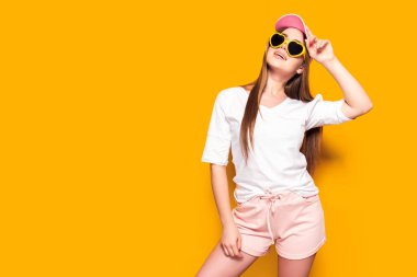 Zarif kıyafetli çekici genç bir bayan şapkaya dokunuyor ve sarı arka planda dikilirken yukarı bakıyor.