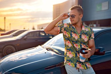 Sonbahar parkında aracın yanındaki tablette elinde bir fincan kahve ve mesaj bloğuyla genç yakışıklı bir adam.