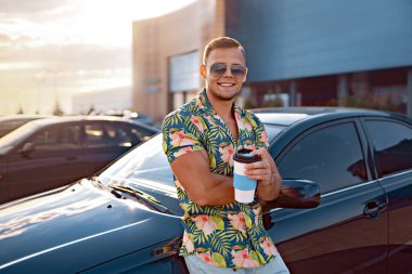 Sonbahar parkında aracın yanındaki tablette elinde bir fincan kahve ve mesaj bloğuyla genç yakışıklı bir adam.