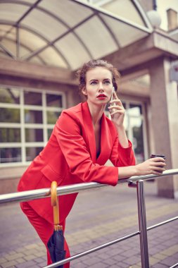Ofis yakınında cep telefonu kullanan kırmızı kostümlü bir iş kadını, cep telefonu takan bir kız, modern banka binasının yakınında akıllı telefondan mesaj atan bir bayan yönetici.