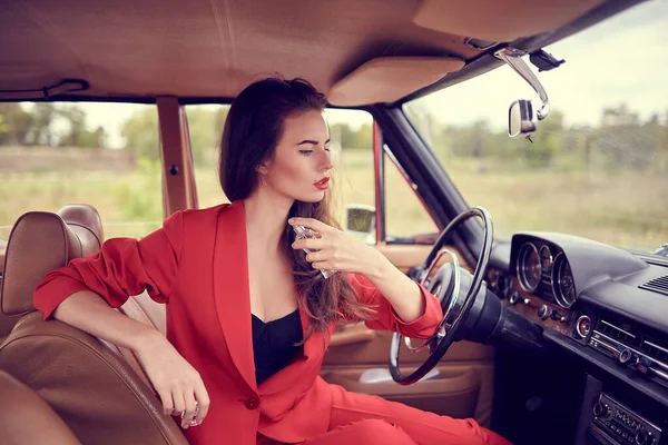 Beautiful young woman in red suit sitting in retro car using a perfume