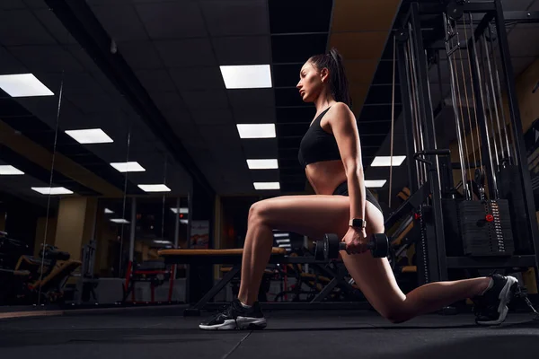 Mujer en forma delgada haciendo embestidas con mancuernas en sus manos en un gimnasio moderno — Foto de Stock