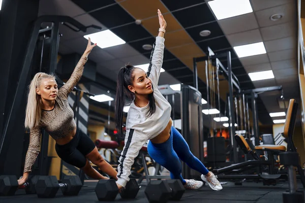 Duas mulheres bonitas com figuras atléticas realizam uma pose de prancha lateral no ginásio. — Fotografia de Stock