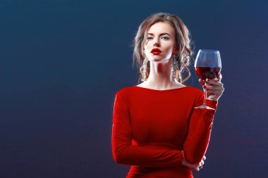 Parlak makyajlı, saç stili, kırmızı elbiseli, koyu arkaplanda bir cam ile poz veren bir kadın.