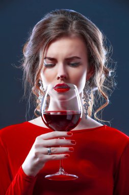 Parlak makyajlı, saç stili, kırmızı elbiseli, koyu arkaplanda bir cam ile poz veren bir kadın.