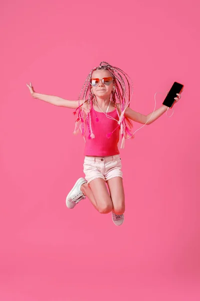 Krásná holčička s dredy skákání s mobilním telefonem v ruce a poslouchat hudbu na sluchátka na růžovém pozadí — Stock fotografie