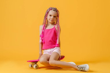 Pembe tişörtlü ve şortlu aktif genç kız kaykayın üzerinde oturuyor.