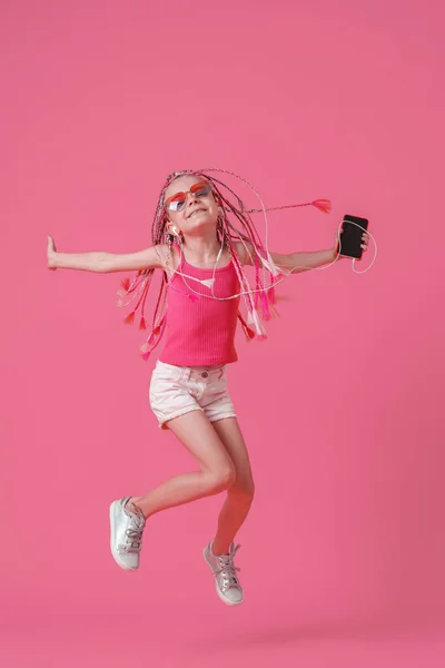 Hermosa niña con rastas saltando con el teléfono móvil en la mano y escuchar música en los auriculares sobre fondo rosa — Foto de Stock