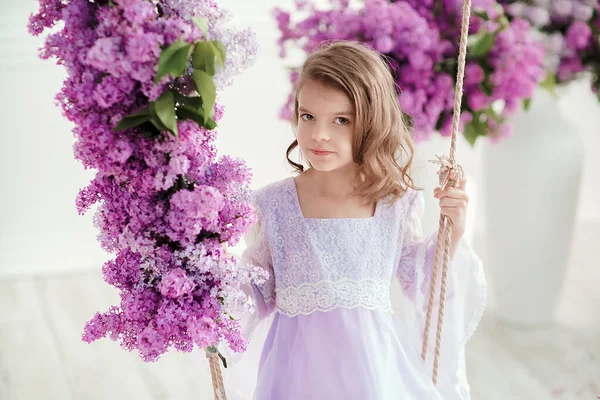 Beautiful little girl of preschool age in a delicate dress sitting on a swing decorated with flowers of lilac. — Stock Photo, Image