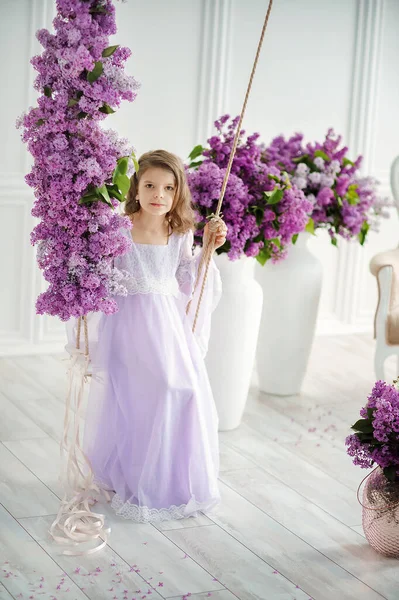Menina bonita da idade pré-escolar em um vestido delicado sentado em um balanço decorado com flores de lilás. — Fotografia de Stock