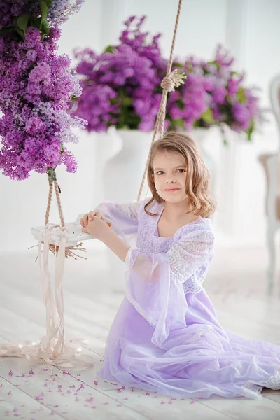 Schöne kleine Mädchen im Vorschulalter in einem zarten Kleid sitzt auf einer Schaukel mit Fliederblumen dekoriert. — Stockfoto