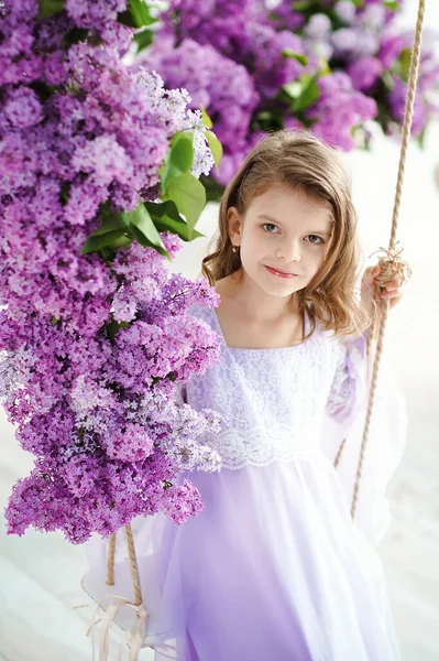 Hermosa niña de edad preescolar en un vestido delicado sentado en un columpio decorado con flores de color lila. — Foto de Stock