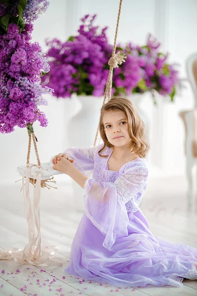 Красивая маленькая девочка дошкольного возраста в нежном платье сидит на качелях, украшенных цветами сирени. — стоковое фото