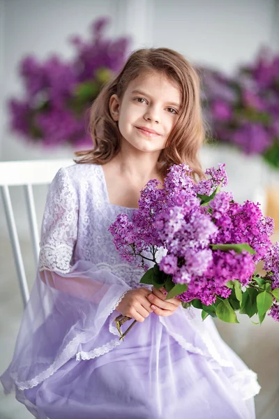 Красивая маленькая девочка дошкольного возраста в нежном платье сидит на качелях, украшенных цветами сирени. — стоковое фото