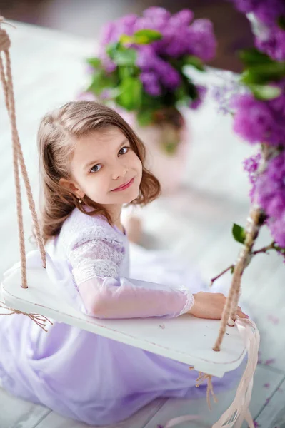Hermosa niña de edad preescolar en un vestido delicado sentado en un columpio decorado con flores de color lila. — Foto de Stock