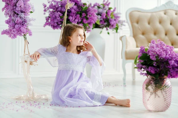 Schöne kleine Mädchen im Vorschulalter in einem zarten Kleid sitzt auf einer Schaukel mit Fliederblumen dekoriert. — Stockfoto