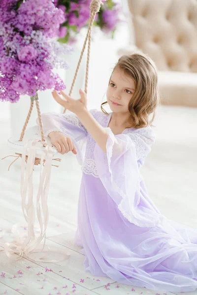 Gyönyörű kislány óvodás korban, finom ruhában, egy hintán ülve, orgonavirágokkal díszítve.. — Stock Fotó