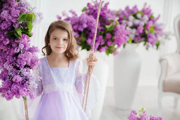 Schöne kleine Mädchen im Vorschulalter in einem zarten Kleid sitzt auf einer Schaukel mit Fliederblumen dekoriert. — Stockfoto