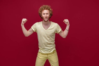 Sarı elbiseli genç adam kırmızı duvar arkasında izole edilmiş bir şekilde duruyor, stüdyo portresi. İnsanlar yaşam tarzı kavramını hisseder. Kopyalama alanı oluşturun. Yükselen eller. Erkeksi duruş