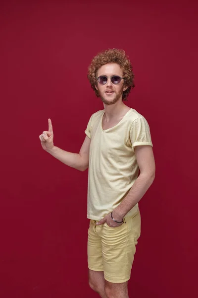Młody mężczyzna z kręconymi włosami w okularach przeciwsłonecznych wskazujący palcem w górę — Zdjęcie stockowe