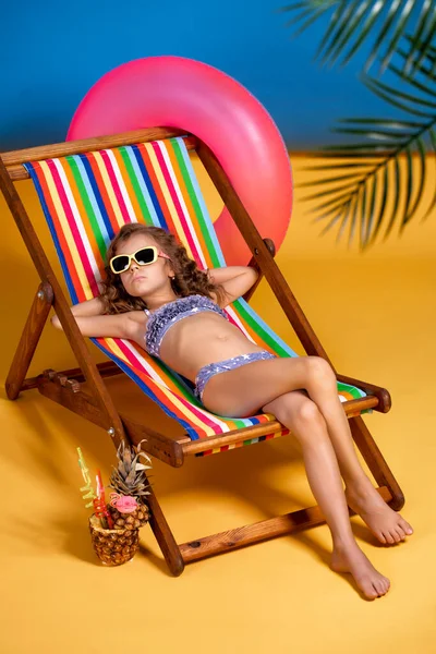 Girl in swimsuit and sunglasses lying in rainbow deck chair with — Stock Photo, Image