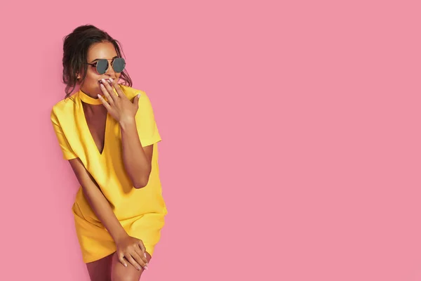Menina bonita em jeans amarelo e camisa rosa, com as mãos para cima usando óculos de sol posando, dançando, sorrindo em fundo rosa no estúdio. — Fotografia de Stock