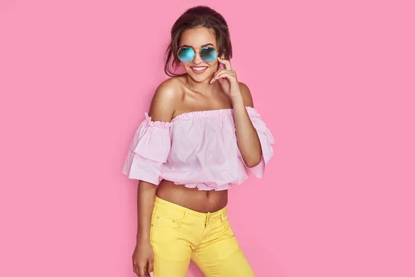 Hermosa chica en jeans amarillos y camisa rosa, con las manos arriba con gafas de sol posando, bailando, sonriendo sobre fondo rosa en el estudio. — Foto de Stock