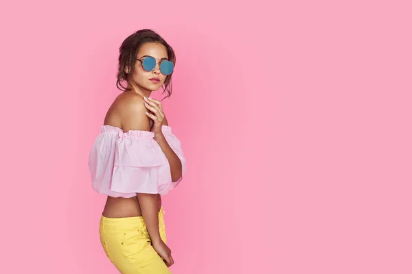 Девушка в желтых джинсах и розовой рубашке, с поднятыми руками в солнцезащитных очках позирует, танцует, улыбается на розовом фоне в студии. — стоковое фото