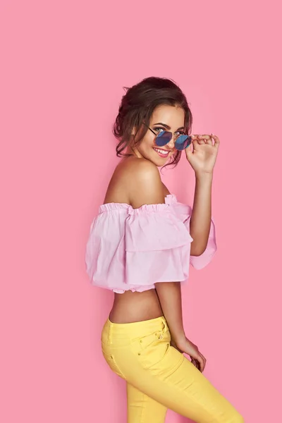 Hermosa chica en jeans amarillos y camisa rosa, con las manos arriba con gafas de sol posando, bailando, sonriendo sobre fondo rosa en el estudio. — Foto de Stock