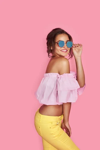 Hermosa chica en jeans amarillos y camisa rosa, con las manos arriba con gafas de sol posando, bailando, sonriendo sobre fondo rosa en el estudio. — Foto de Stock
