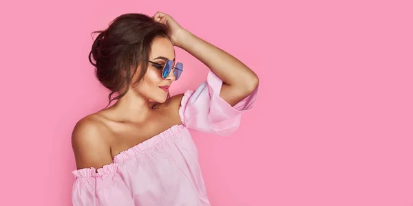 Hermosa chica en jeans amarillos y camisa rosa, con las manos arriba con gafas de sol posando, bailando, sonriendo sobre fondo rosa en el estudio. — Foto de Stock