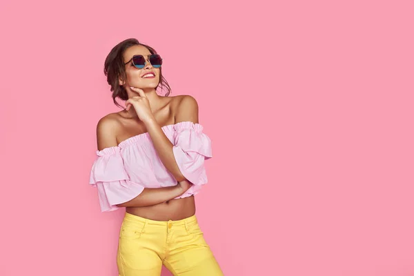 Hermosa chica en jeans amarillos y camisa rosa, con las manos arriba con gafas de sol posando, bailando, sonriendo sobre fondo rosa en el estudio. — Foto de Stock