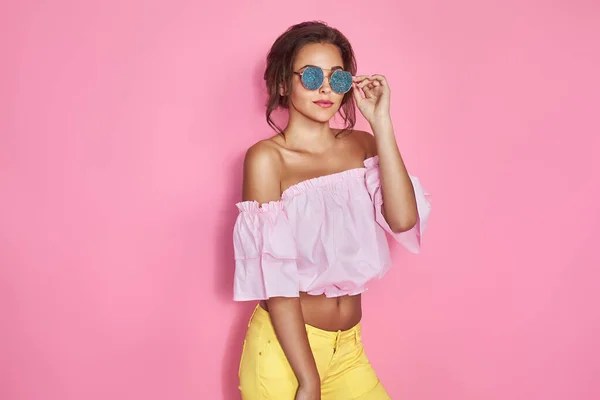 Hermosa chica en jeans amarillos y camisa rosa, con las manos arriba con gafas de sol posando, bailando, sonriendo sobre fondo rosa en el estudio. — Foto de Stock