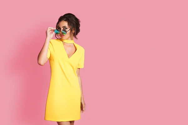 Hermosa chica en vestido amarillo con gafas de sol posando, sonriendo sobre fondo rosa en el estudio. —  Fotos de Stock