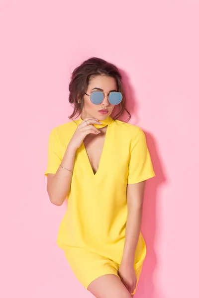 Красива дівчина в жовтій сукні в сонцезахисних окулярах позує, посміхається на рожевому фоні в студії . — стокове фото