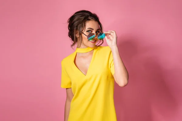 Gadis cantik dengan gaun kuning berpose kacamata hitam, tersenyum di latar belakang merah muda di studio. — Stok Foto