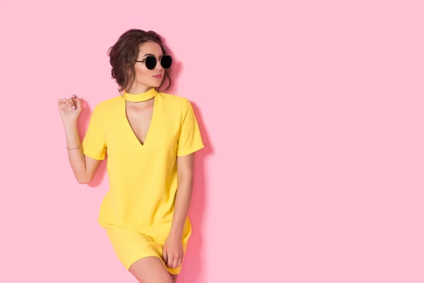 Gadis cantik dengan gaun kuning berpose kacamata hitam, tersenyum di latar belakang merah muda di studio. — Stok Foto