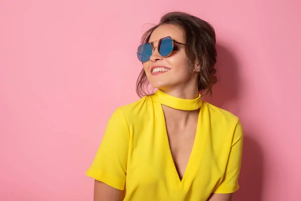 Hermosa chica en vestido amarillo con gafas de sol posando, sonriendo sobre fondo rosa en el estudio. —  Fotos de Stock