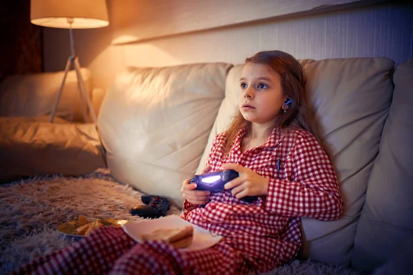 Happy little girl in pajams sitting on sofa, eating pizza and pl — Stock Photo, Image