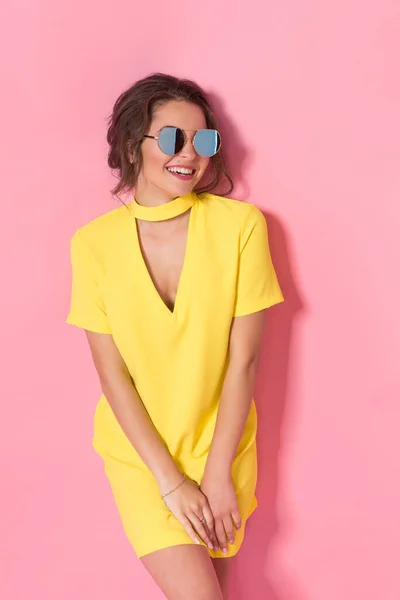 Gyönyörű lány sárga ruha visel napszemüveg pózol, mosolygós rózsaszín háttér stúdió. — Stock Fotó
