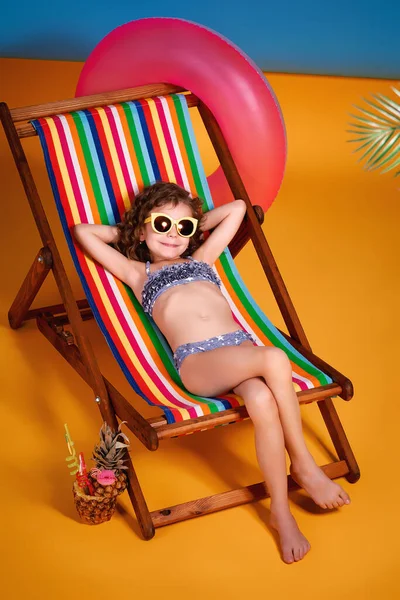 Smiling girl in lilac swimsuit and sunglasses lying in rainbow deck chair with crossed legs and sunbathing — Stock Photo, Image