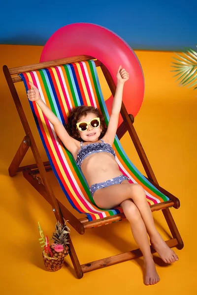 Uśmiechnięta dziewczyna w liliowym stroju kąpielowym i okularach przeciwsłonecznych leżąca w tęczowym fotelu z skrzyżowanymi nogami i opalaniem — Zdjęcie stockowe