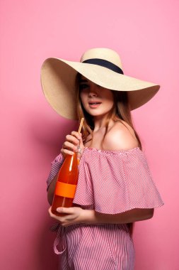 Çekici bir kadının hasır şapka giyip pembe arka planda izole edilmiş cam şişeden meyve suyu içtiği fotoğraf.. 