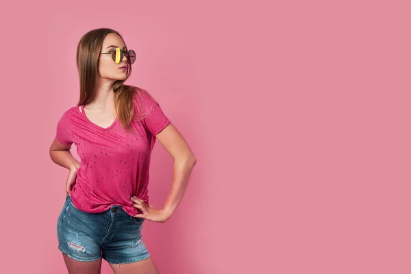 Фотография красивой молодой женщины на розовом фоне в розовой футболке и стильных красочных солнцезащитных очках, смотрящих в камеру — стоковое фото
