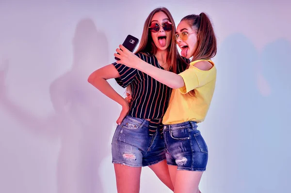 Twee tienermeisjes in zomerkleren staan in de studio en maken selfie op witte achtergrond. Gemengde verlichting — Stockfoto