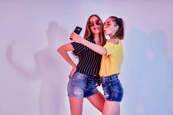 Dvě dospívající dívky v letním oblečení stojící ve studiu a dělat selfie na bílém pozadí. Smíšené světlo — Stock fotografie