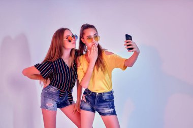 Stüdyoda duran ve beyaz arka planda selfie çeken iki genç kız. Karışık ışık